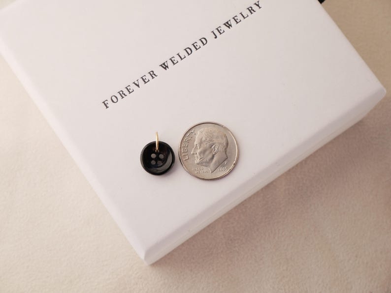 May include: A black button pendant with a gold-colored bail next to a dime on a white box. The button has four holes and a shiny finish. The box has the words "FOREVER WELDED JEWELRY" printed on it.