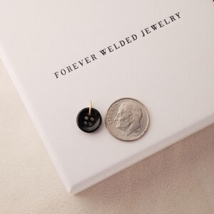 May include: A small, black button pendant with a gold-colored loop next to a dime on a white surface. The button has four holes. The text "FOREVER WELDED JEWELRY" is printed on the white surface.