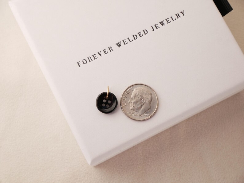May include: A small, black button pendant with a gold-colored loop next to a dime on a white surface. The button has four holes. The text "FOREVER WELDED JEWELRY" is printed on the white surface.
