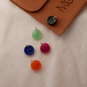 May include: A collection of colorful button charms in green, blue, red, and orange, each with a gold-colored loop. A black button charm and a brown leather pouch are also present. The buttons are on a white surface.