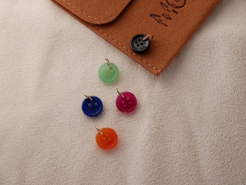 May include: A collection of colorful button charms in green, blue, red, and orange, each with a gold-colored loop. A black button charm and a brown leather pouch are also present. The buttons are on a white surface.