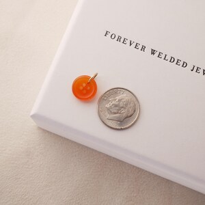 May include: An orange button pendant with a gold bail sits next to a US dime on a white surface. The pendant is round with four small holes. The text "FOREVER WELDED JEWELRY" is printed on the white surface.
