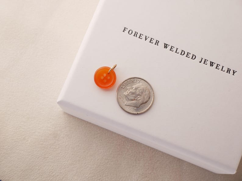 May include: An orange button pendant with a gold bail sits next to a US dime on a white surface. The pendant is round with four small holes. The text "FOREVER WELDED JEWELRY" is printed on the white surface.