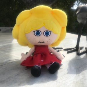Könnte beinhalten: Eine Plüschpuppe mit blonden Haaren, die ein rotes Kleid mit weißen Punkten trägt. Die Puppe hat blaue Augen und ein lächelndes Gesicht.