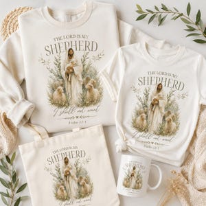 Puede incluir: Sudadera, camiseta, bolso de mano y taza de color crema con el texto "The Lord is My Shepherd" y una ilustración de Jesús y ovejas. El diseño incluye el texto "I shall not want" y Salmo 23:1.
