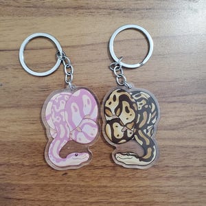 May include: Two keychains with snake designs. One keychain features a pink snake with white patterns, and the other has a brown and tan snake design. Both keychains have silver key rings and chains.
