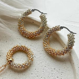 May include: A jewelry set featuring a beaded necklace and hoop earrings. The necklace has a gold chain with a beaded circle pendant. The earrings are hoop style, with a mix of gold and silver beads. The jewelry is displayed on a white surface.