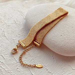 Puede incluir: Un brazalete de cadena dorado con un diseño tejido y un cierre de langosta. El brazalete tiene una pequeña etiqueta que dice "S.Steel".