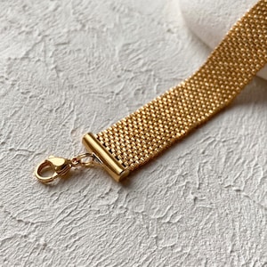 May include: A gold bracelet featuring a woven mesh design. The bracelet has a rectangular shape and a clasp closure. The metal has a shiny finish. The bracelet is shown against a textured white background.