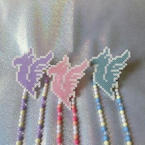 May include: Three beaded necklaces with unicorn-shaped charms. The charms are made of colorful perler beads in purple, pink, and blue. The necklaces are made of white beads with accents of purple, pink, and blue beads.