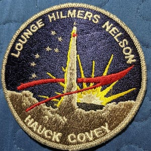 Könnte beinhalten: Ein runder, gestickter Aufnäher mit dunkelblauem Hintergrund. Der Aufnäher zeigt einen stilisierten Raketenstart mit einer roten und gelben Flamme. Der Text "LOUNGE HILMERS NELSON" ist kreisförmig um den oberen Rand des Aufnähers geschrieben. Der Text "HAUCK COVEY" ist kreisförmig um den unteren Rand des Aufnähers geschrieben.