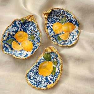 May include: Three decorative oyster shells featuring a blue and white tile pattern with yellow lemon illustrations and green leaves. Each shell has a metallic gold painted edge, creating a distinct contrast against the intricate floral motifs.