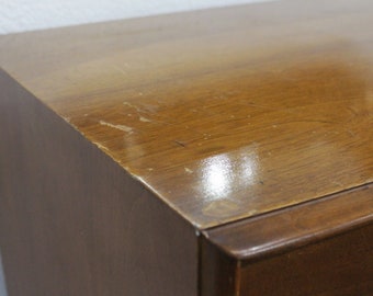 Può includere: Primo piano della parte superiore di una cassettiera in legno con finitura marrone chiaro. Il legno ha una leggera lucentezza e ci sono alcuni graffi sulla superficie.