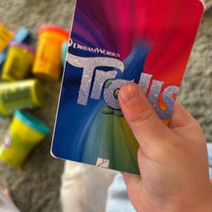May include: A close-up of a card with a colourful background and the DreamWorks Trolls logo. The card is being held by a hand.