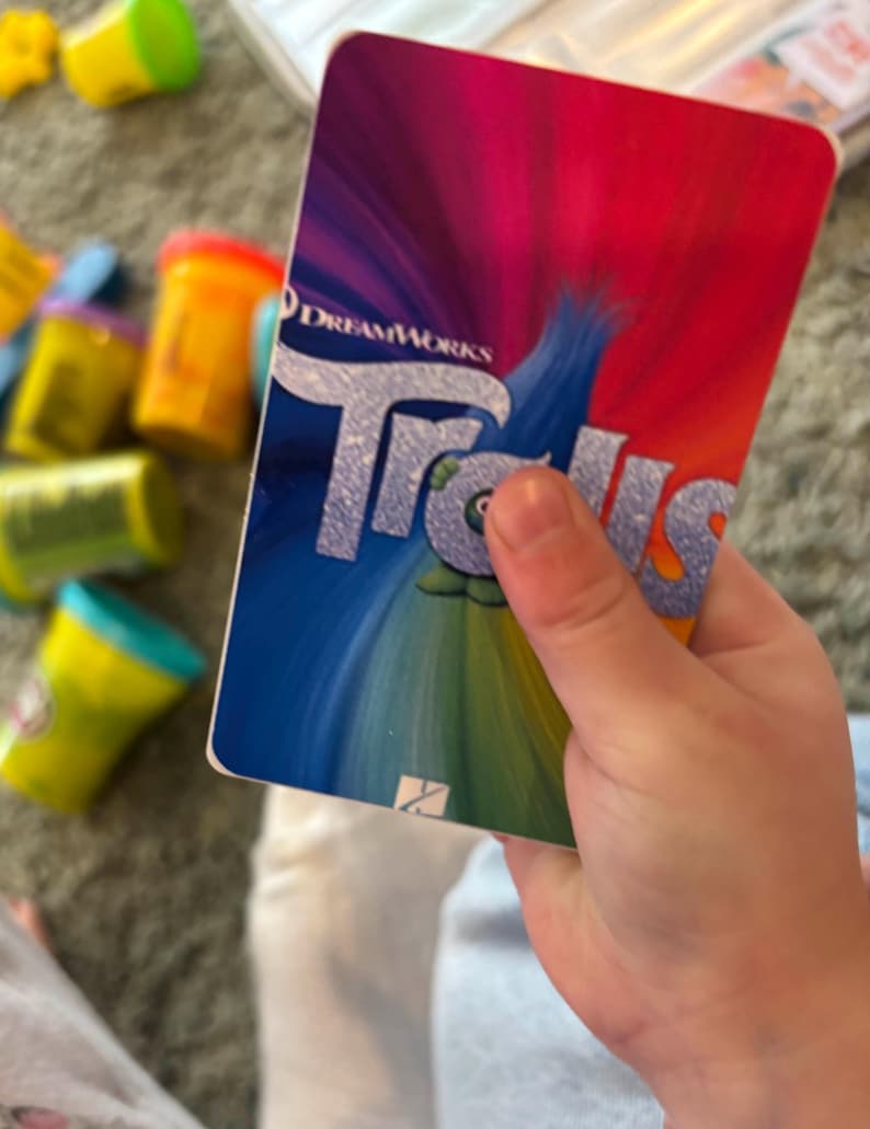May include: A close-up of a card with a colourful background and the DreamWorks Trolls logo. The card is being held by a hand.