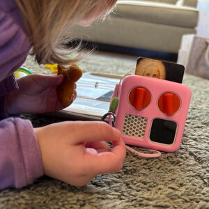 May include: A pink toy with two red buttons, a black screen, and a white honeycomb pattern. The toy is on a grey carpet.