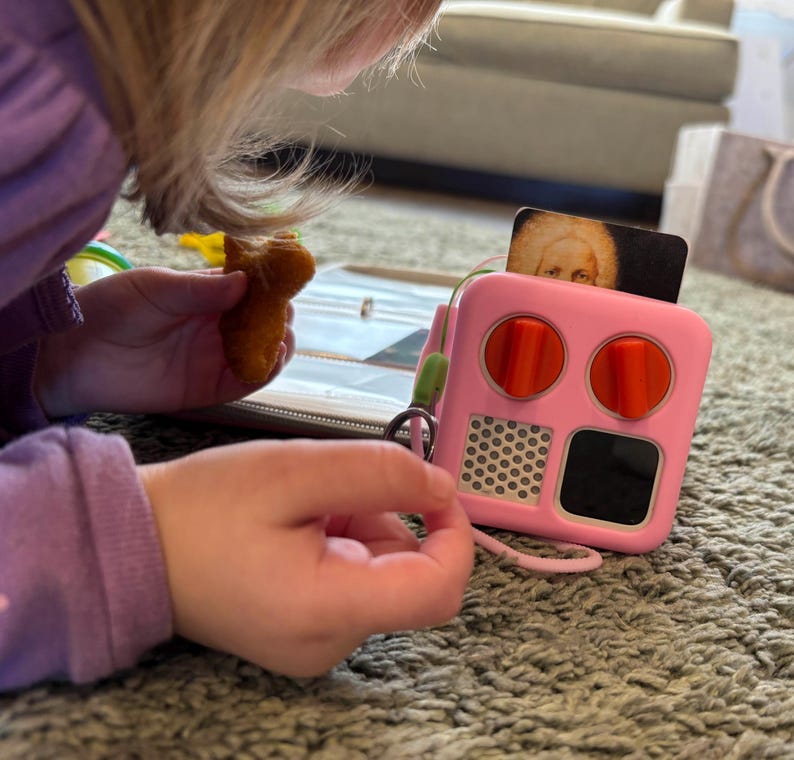 May include: A pink toy with two red buttons, a black screen, and a white honeycomb pattern. The toy is on a grey carpet.