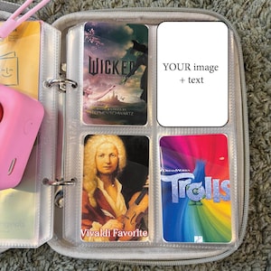 May include: A clear plastic binder containing a selection of colourful cards. The cards feature images of a musical score, a portrait of a man, a movie poster, and a musical play. The binder has a ring mechanism for holding the cards.