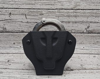 Könnte beinhalten: Ein schwarzes Lederholster für Handschellen mit einer silbernen Handschelle im Inneren. Das Holster hat eine strukturierte Oberfläche und zwei kleine Schrauben an der Seite.