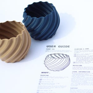 A navy blue and beige coloured 3D printed plant pot sitting next to a user guide, photographed on a white background.