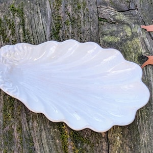 Peut inclure: Un plat de service en céramique blanche en forme de coquille avec un bord cannelé. Le plat est posé sur une surface en bois.