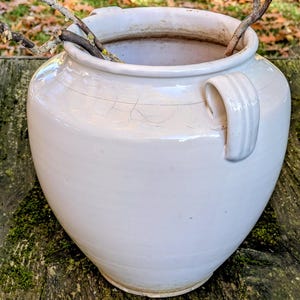 Op de afbeelding: Een grote, crèmekleurige keramische vaas met een afgeronde body en een enkele handgreep. De vaas heeft zichtbare scheuren en bevat droge takken. De achtergrond toont een houten oppervlak en onscherpe bladeren.