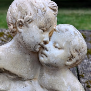 Peut inclure: Une sculpture en pierre blanc cassé patinée représentant deux enfants aux visages rapprochés. Les personnages présentent des surfaces texturées et vieillies avec des signes d'usure visibles, montrant des traits détaillés.