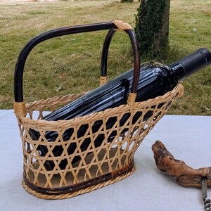 Peut inclure: Un porte-bouteille en osier tressé avec une anse arquée marron foncé et un corps en treillis beige clair, contenant une bouteille de vin en verre sombre. À côté du support se trouve un tire-bouchon en bois rustique avec un mécanisme en spirale.