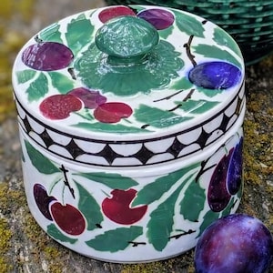 Può includere: Una brocca in ceramica verde scuro con un motivo a intreccio testurizzato e un barattolo in ceramica abbinato con coperchio, decorato con foglie verdi dipinte e prugne rosse e viola, accompagnato da tre prugne viola fresche intere.