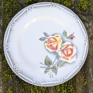 May include: White ceramic plate with a decorative border and a floral design featuring two yellow roses with red centers. The roses are painted on a stem with green leaves.