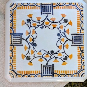 May include: Square ceramic tile with a white background, featuring a floral design in orange, black, and gray. The tile has a decorative border and four black rectangular accents. The floral pattern includes orange bell-shaped flowers and black stems.