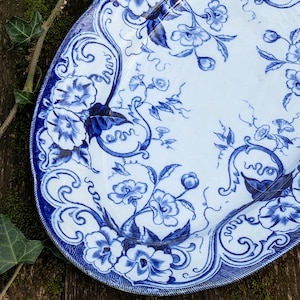 Peut inclure: Un plat ovale en céramique à fond blanc orné d'un motif floral bleu. Le design présente des illustrations détaillées de fleurs, de feuilles et de vignes entrelacées, avec une bordure décorative dotée d'une finition bleue texturée.