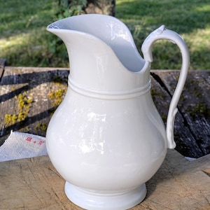 Può includere: Una brocca in ceramica bianca con finitura lucida, caratterizzata da un corpo arrotondato, un ampio beccuccio e un manico curvo. La brocca presenta una fascia orizzontale in rilievo attorno al collo e poggia su una base circolare liscia.