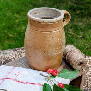 Puede incluir: Una jarra de cerámica rústica de color marrón claro con asa, colocada sobre una superficie de madera. También hay un rollo de cuerda, una ramita de cerezas y un paño de lino a rayas con bordados rojos. La jarra tiene una abertura ancha y una superficie texturizada.