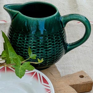 Op de afbeelding: Een groene keramische kan met een geweven patroon en een handvat. De kan staat op een houten snijplank naast een wit bord met een rood en wit ontwerp.