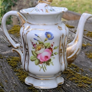 Può includere: Una teiera in porcellana bianca con un bouquet floreale composto da una rosa rosa, un fiore di gloria mattutina blu e accenti gialli. La teiera presenta un manico curvo, un beccuccio e un coperchio con finiture dorate lungo i bordi e i pannelli.