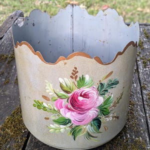 Peut inclure: Un récipient cylindrique en métal avec un bord supérieur festonné, présentant une finition beige craquelée et un motif floral peint à la main. Le motif central montre une grande rose rose entourée de feuilles vertes et de brindilles brunes.