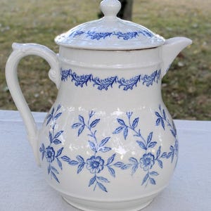 Pode incluir: Um jarro de cerâmica branca com tampa, apresentando um padrão floral azul. O design inclui um delicado motivo de videiras e flores ao redor do corpo e uma borda decorativa na tampa. Possui uma alça curva e um pequeno bico para servir.