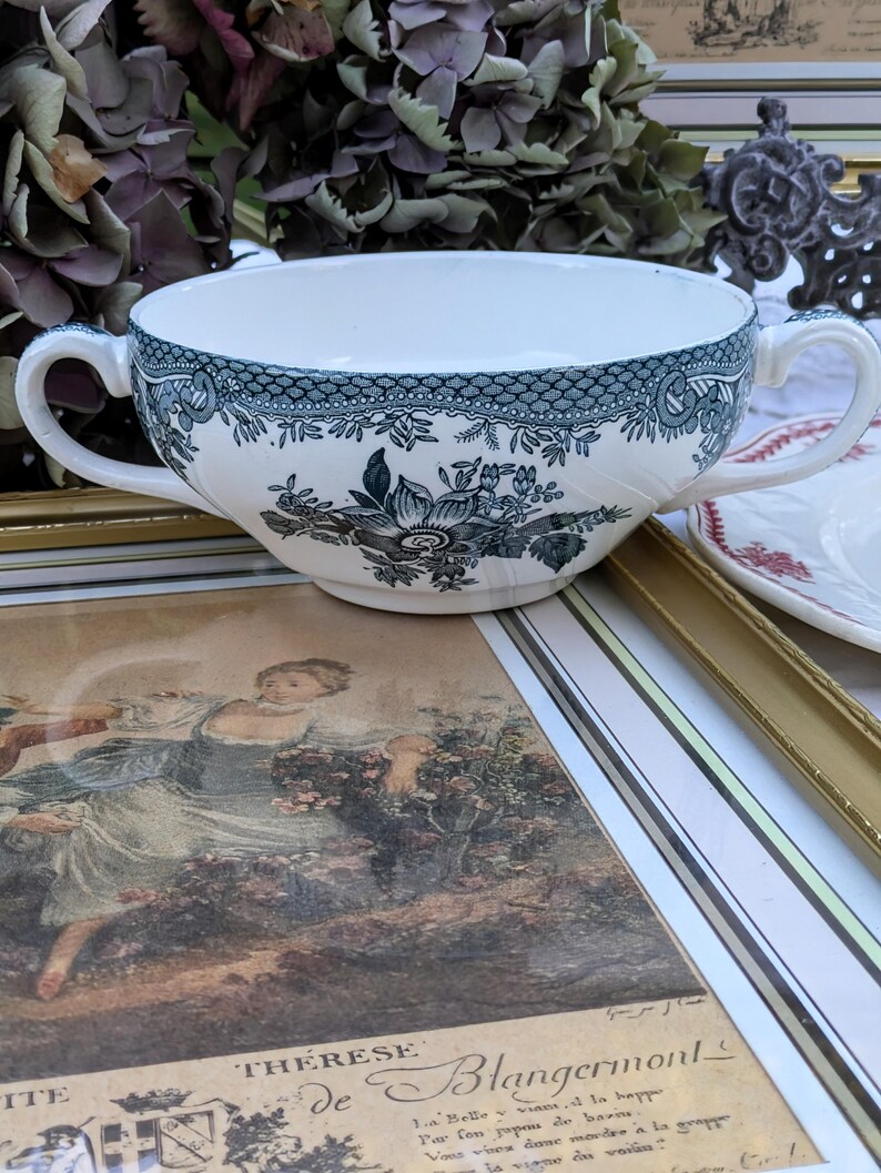 K&ouml;nnte beinhalten: Eine wei&szlig;e Suppensch&uuml;ssel aus Keramik mit einem blauen und gr&uuml;nen Blumenmuster. Die Sch&uuml;ssel hat zwei Henkel und einen gewellten Rand.