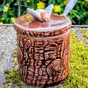 Op de afbeelding: Een bruine keramische honingpot in de vorm van een boomstromp met reliëf van bijenfiguren aan de zijkanten. Het deksel heeft een gebeeldhouwde bij als handvat en een zilverkleurige metalen lepel die door een kleine inkeping steekt.