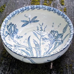 Op de afbeelding: Een ronde keramische kom met een crèmekleurige basis voorzien van blauwe botanische illustraties van irissen en een libel. De rand is versierd met een herhalend blauw bloemenpatroon en de binnenzijde toont gedetailleerde blauwe bloemstengels.