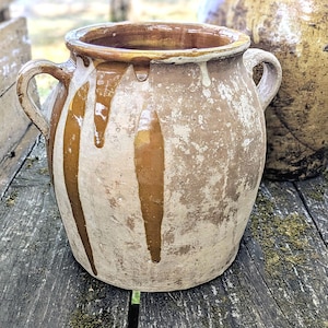 May include: A rustic earthenware pot with two side handles, featuring a light beige body with vertical brown glaze drips running from the rim. The surface has a weathered, textured appearance with visible signs of age and a wide, open circular mouth.