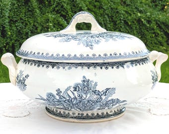 Op de afbeelding: Een crèmekleurige keramische terrine met bijpassend deksel, voorzien van een donkerblauw bloemenpatroon. De schaal heeft twee handvatten aan de zijkanten en een decoratieve knop op het deksel, met een geometrische rand rondom de voet en de rand.