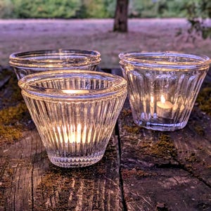 May include: Three clear glass votive candle holders with a vertical ribbed texture. Each holder features a wide, flared rim and a tapered base. Two of the holders contain lit tea light candles, casting a warm glow through the transparent glass.
