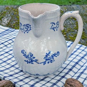 Op de afbeelding: Een crèmekleurige keramische kan met een zichtbare craquelé glazuurafwerking, voorzien van blauwe bloemmotieven rondom het lichaam en de rand. De kan heeft een gebogen handvat en een schenktuit, met een vintage uitstraling en gedetailleerde blauwe botanische motieven.