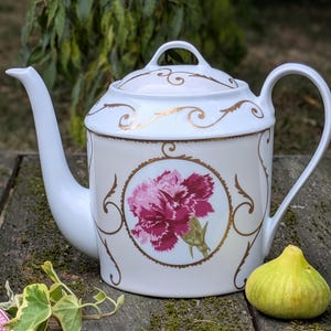 Op de afbeelding: Witte porseleinen theepot met deksel, tuit en handvat. De theepot heeft een roze en rode bloemdesign in een goudkleurig frame. Gouden krullen versieren het deksel en de body. Een groene vijg ligt in de buurt.