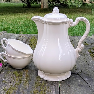 May include: A cream-colored porcelain coffee pot with a lid, two teacups, and a handle. The coffee pot has a bulbous body and a decorative handle. The teacups are stacked and have a curved handle. The set is on a weathered wooden surface.