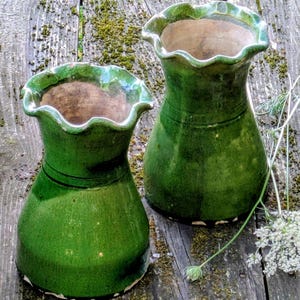 Puede incluir: Dos jarrones de cerámica verdes con bordes ondulados. Los jarrones tienen un acabado brillante y una base bulbosa. El interior de los jarrones es de un color marrón contrastante. Los jarrones se exhiben sobre una superficie de madera envejecida.