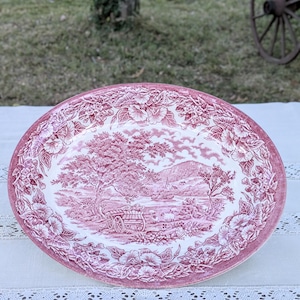 Puede incluir: Una bandeja ovalada de cerámica con un patrón de transferencia en rosa y blanco. El centro representa un paisaje rural con una carreta tirada por caballos, árboles y ovejas. El borde ancho está decorado con un motivo floral detallado.