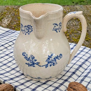 Peut inclure: Un pichet en céramique blanche avec un motif floral bleu. Le pichet a une anse et un bec verseur. Il est posé sur une nappe à carreaux bleus et blancs.